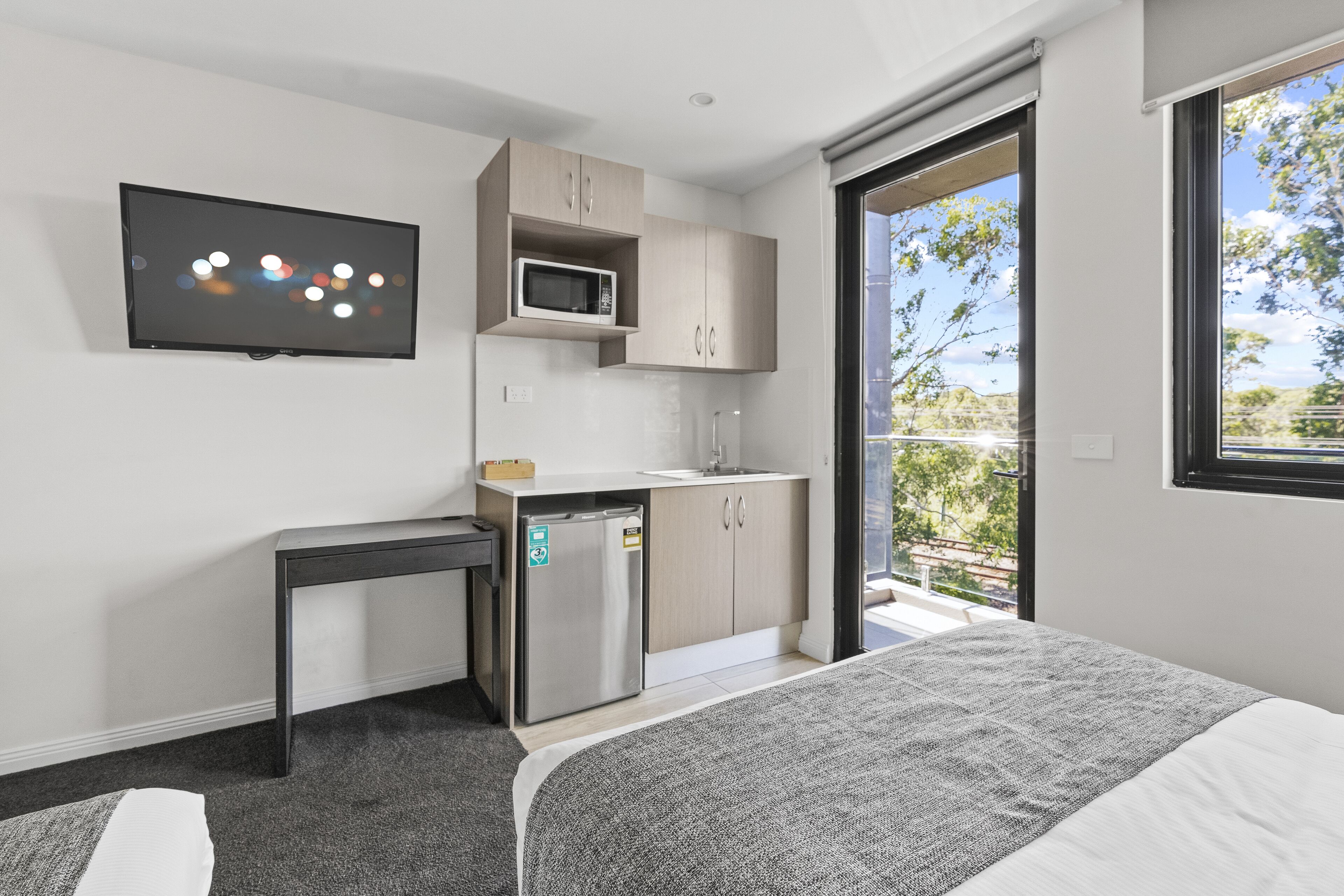 A modern bedroom with a bed, a small kitchenette, a flat-screen TV, and a balcony with a view.