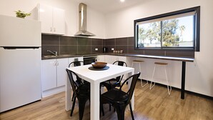 A modern kitchen with white cabinetry, a stainless steel range hood, a dining table with four chairs, and a window with a view of trees.