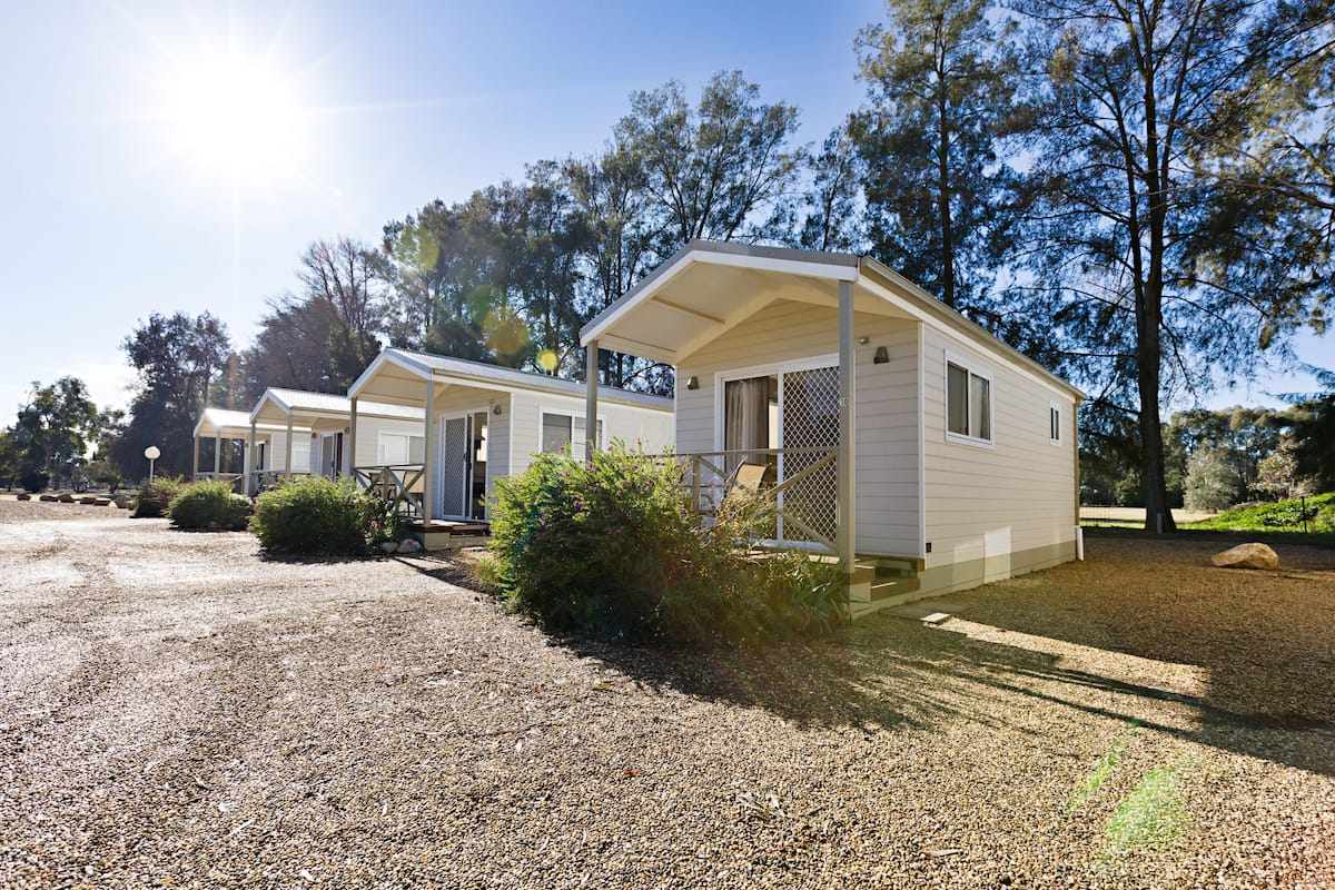 A row of single-story mobile homes with attached porches, surrounded by gravel and trees.