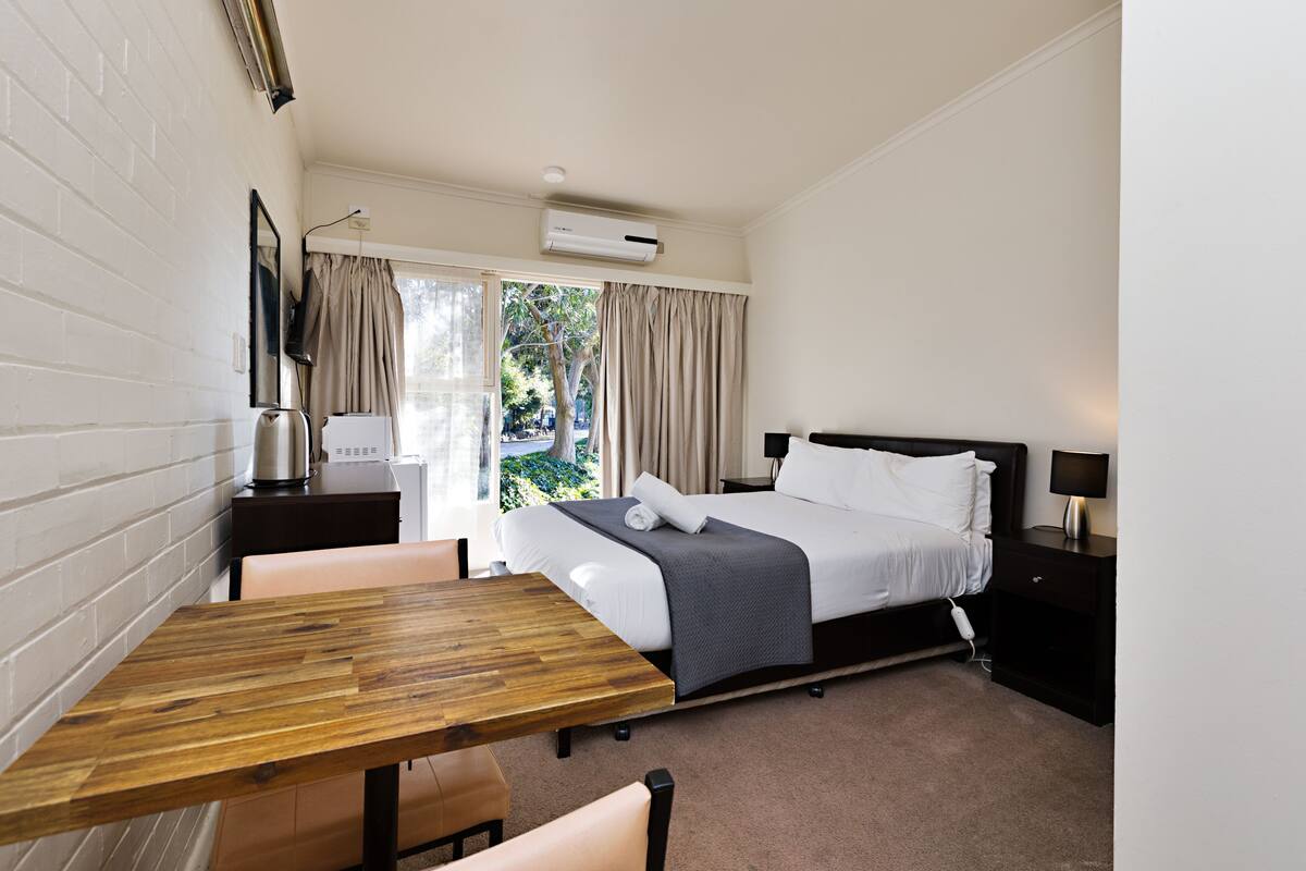 A hotel room with a bed, bedside table, lamp, and a view of the outdoors.