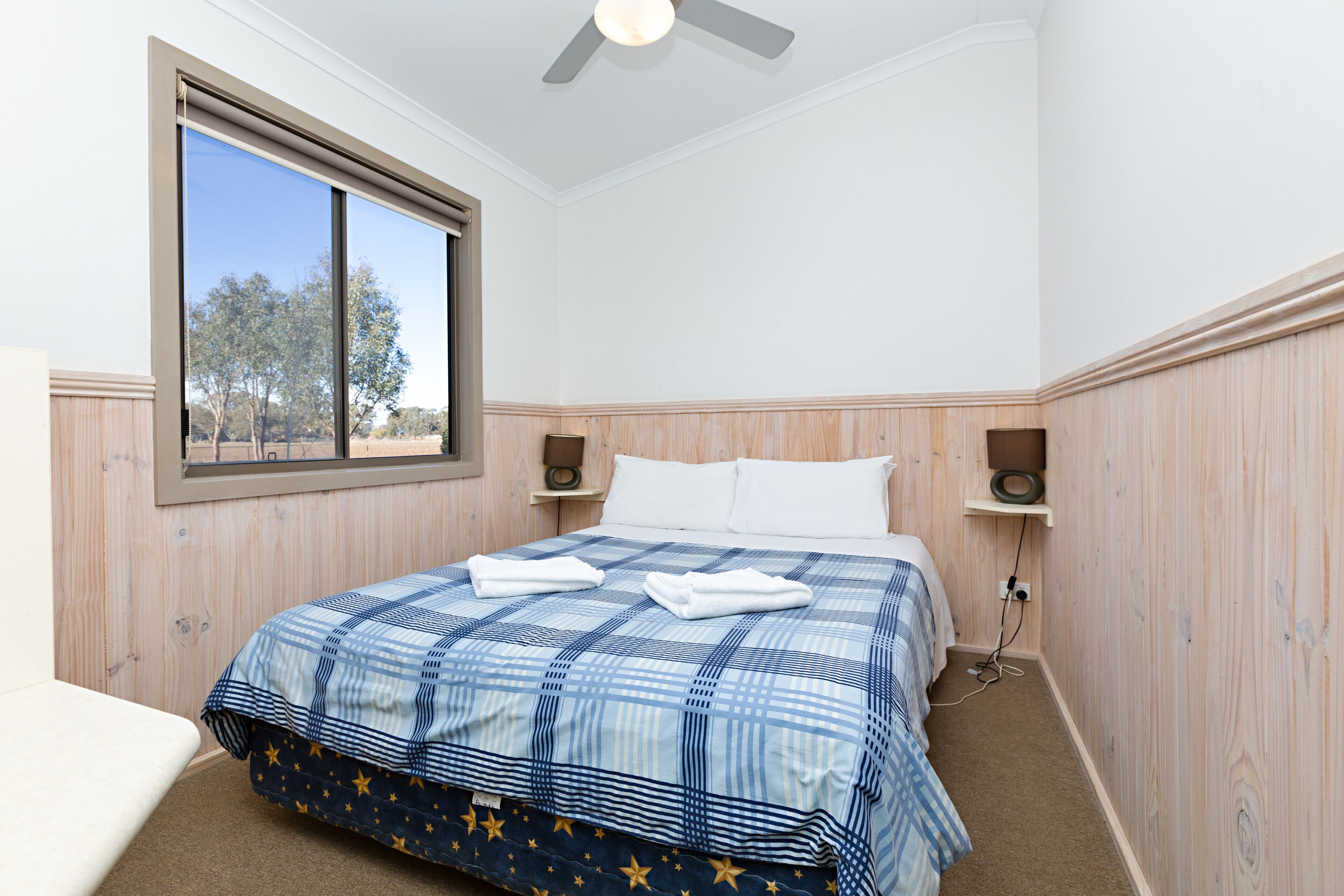 A bedroom with a large bed, a window with a view of trees, wooden paneling, and a ceiling fan.