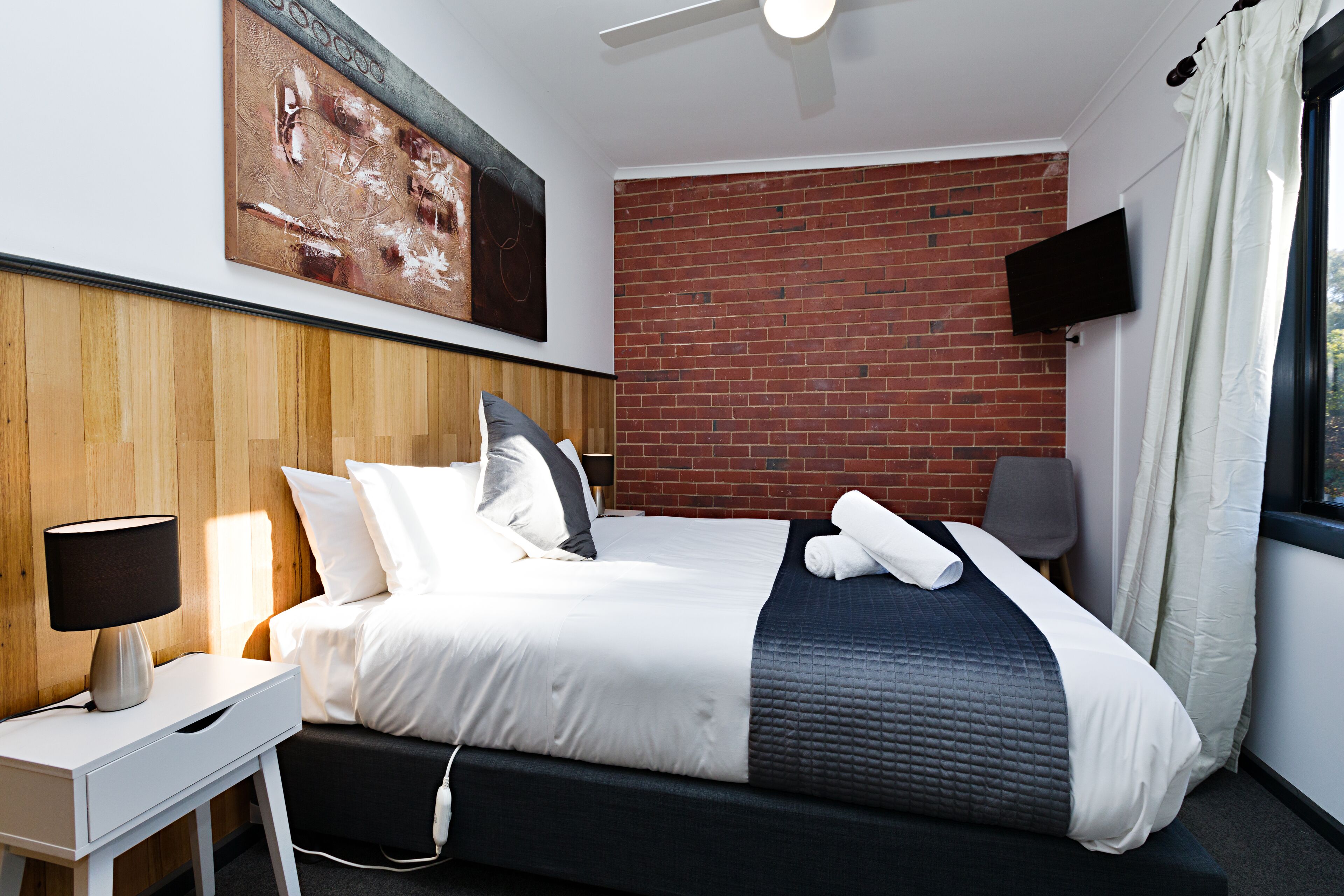 Ein modernes Hotelzimmer mit Backsteinwand, Flachbildfernseher, Nachttisch mit Lampe und einem ordentlich bezogenen Bett.
