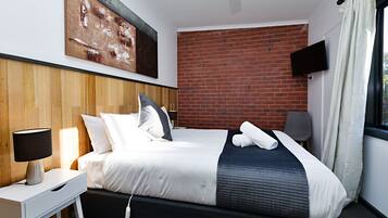A modern hotel room with a brick wall, a flat-screen TV, a bedside table with a lamp, and a neatly made bed.