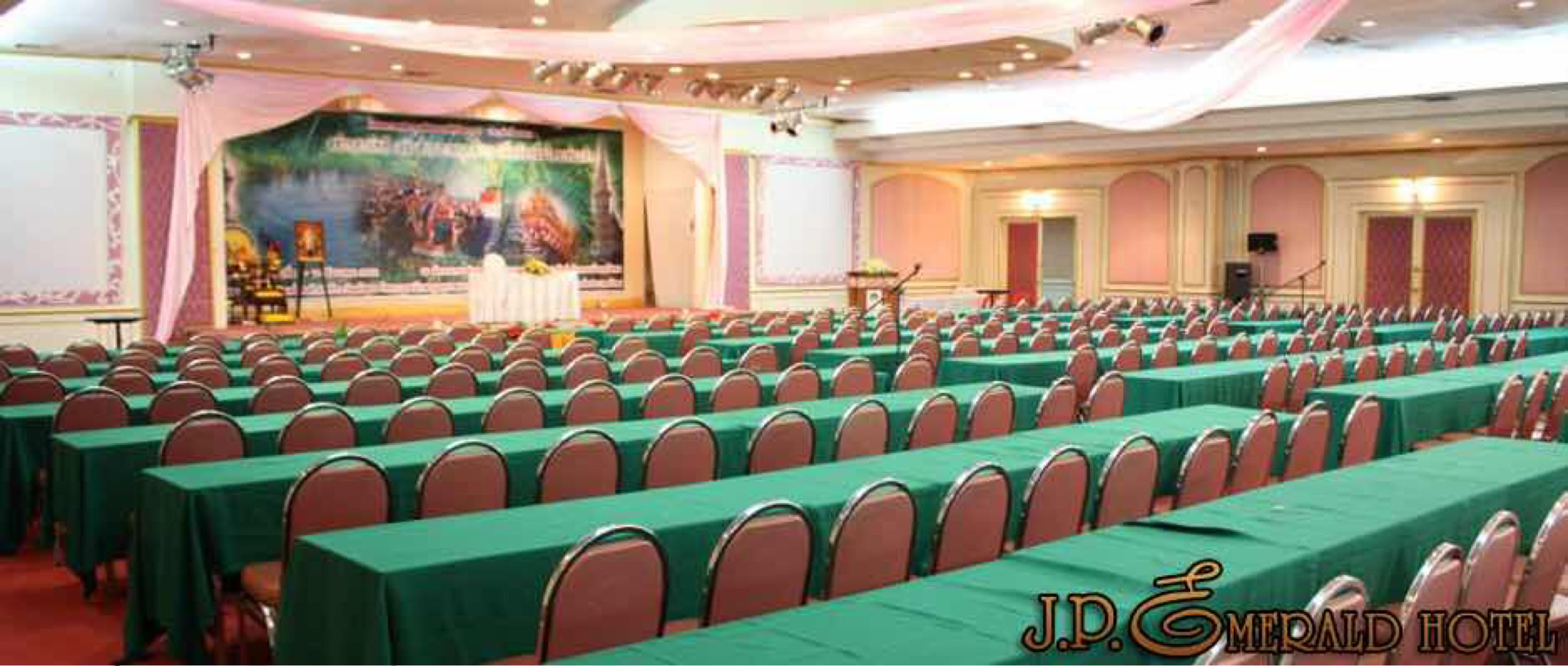 Un salón de conferencias con filas de mesas y sillas, un escenario con fondo decorativo y un atril para oradores.