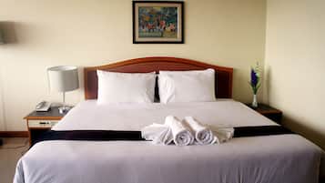A neatly made bed with white linens, a wooden headboard, a bedside table with a lamp, a phone, and a vase with flowers.