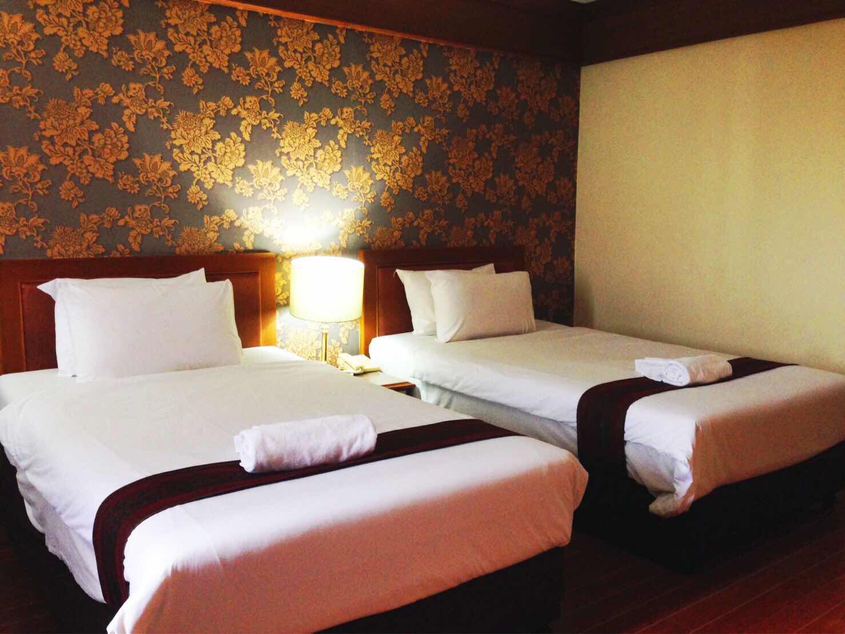 Habitación de hotel con dos camas, cabecera de madera, mesita de noche con lámpara y papel tapiz con motivos florales.