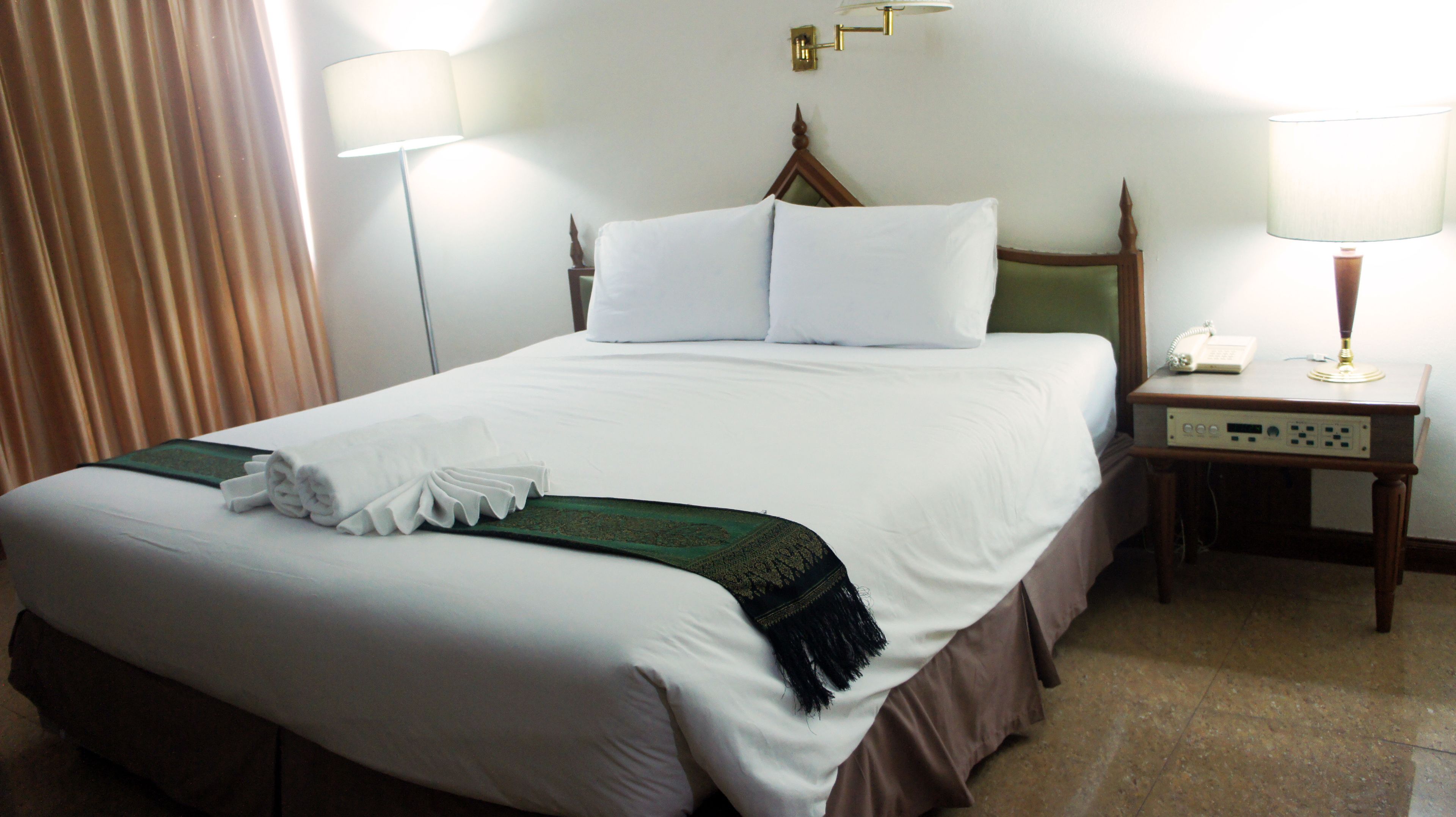 Una cama bien hecha con sábanas blancas, un plaid verde y una toalla blanca doblada. Hay una mesita de noche de madera con una lámpara y un teléfono. La habitación tiene un diseño sencillo y limpio con una cortina a la izquierda.