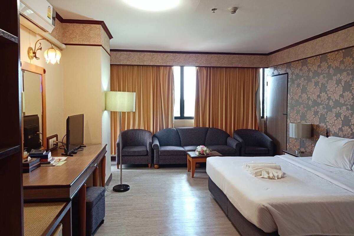 A hotel room with a bed, two armchairs, a desk with a computer, a lamp, and a window with curtains.