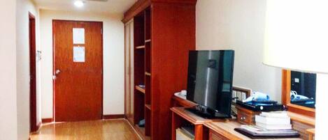 A hotel room with a wooden floor, a desk with a television, a mini-fridge, and a wardrobe.