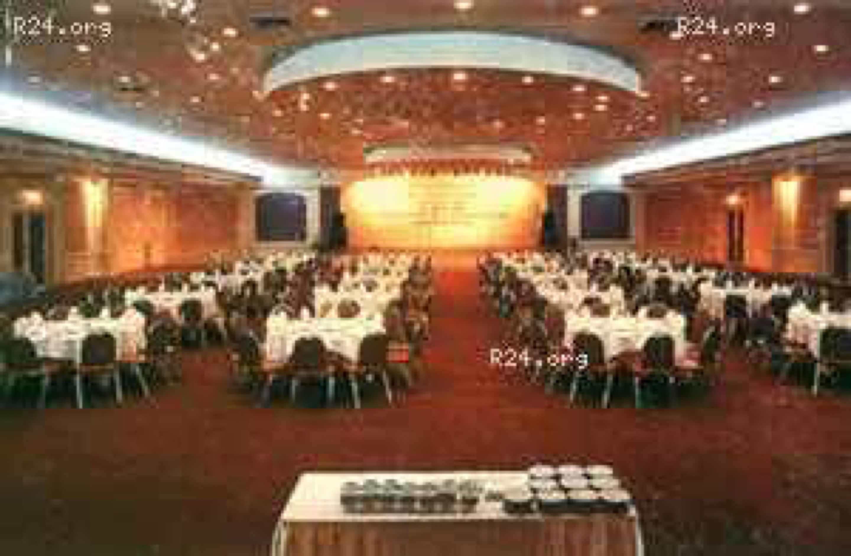 A large conference room with rows of tables and chairs, a stage at the front, and a high ceiling with recessed lighting.