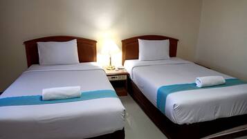 A hotel room with two single beds, each with a white bedspread and a blue accent. There is a nightstand with a lamp between the beds.