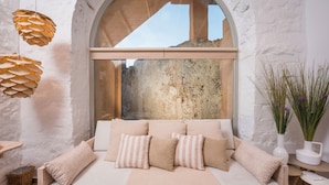 Una habitación acogedora con un sofá blanco, almohadones estampados y un ventanal con vista a una pared rústica.