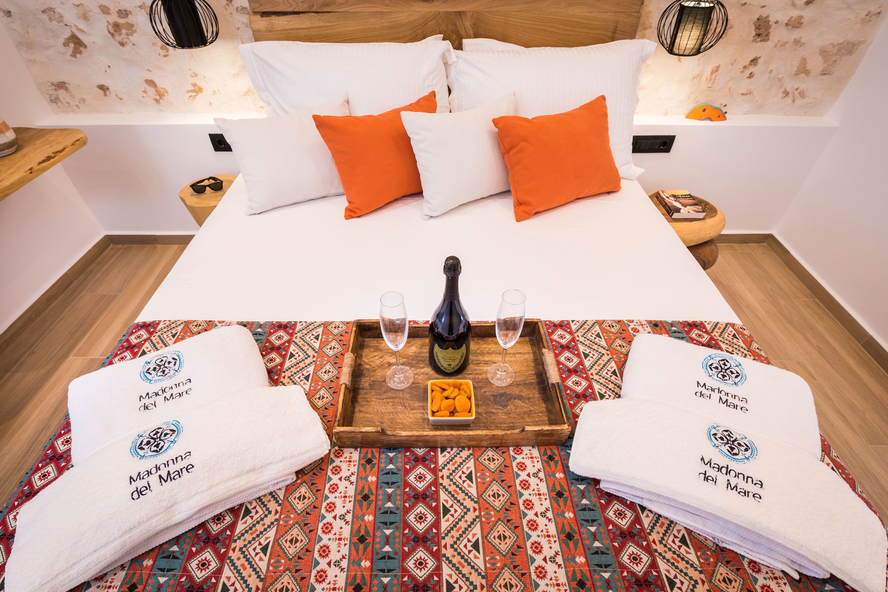 Una cama bien tendida con almohadas blancas y naranjas, una bandeja de madera con una botella y vasos, y dos toallas con un logotipo.