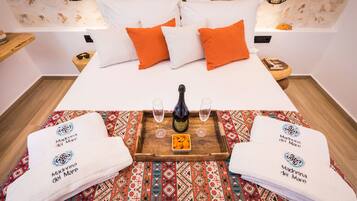 Una cama bien tendida con almohadas blancas y naranjas, una bandeja de madera con una botella y vasos, y dos toallas con un logotipo.