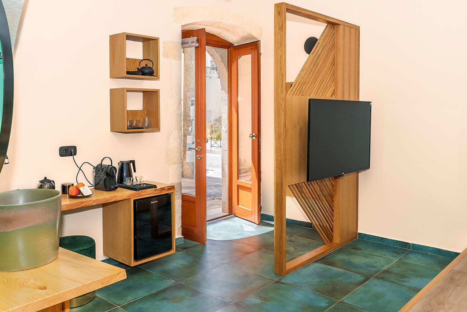 Una cocina compacta con cafetera, hervidor y un refrigerador pequeño. Un mueble de televisión de madera sostiene un televisor de pantalla plana. La habitación tiene piso de baldosas y una puerta de madera que da a un área exterior.