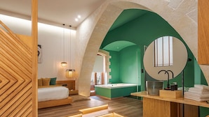 Una habitación de hotel moderna con suelo de madera, cama con ropa de cama blanca, pared verde decorativa, espejo redondo y baño pequeño con bañera.