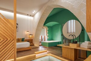 Une chambre d’hôtel moderne avec un plancher en bois, un lit recouvert de linge de maison blanc, un mur d’accent vert, un miroir rond et une petite salle de bain avec une baignoire.