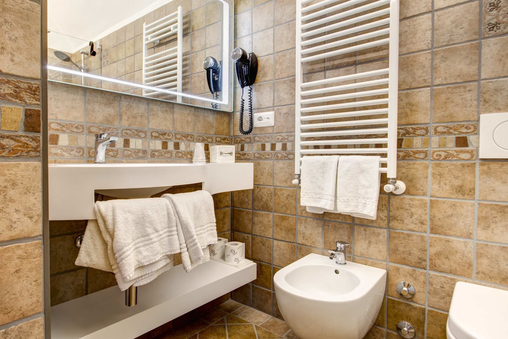 Un baño con lavamanos blanco, bidé y toallero. Hay un espejo sobre el lavamanos y un toallero con toallas.