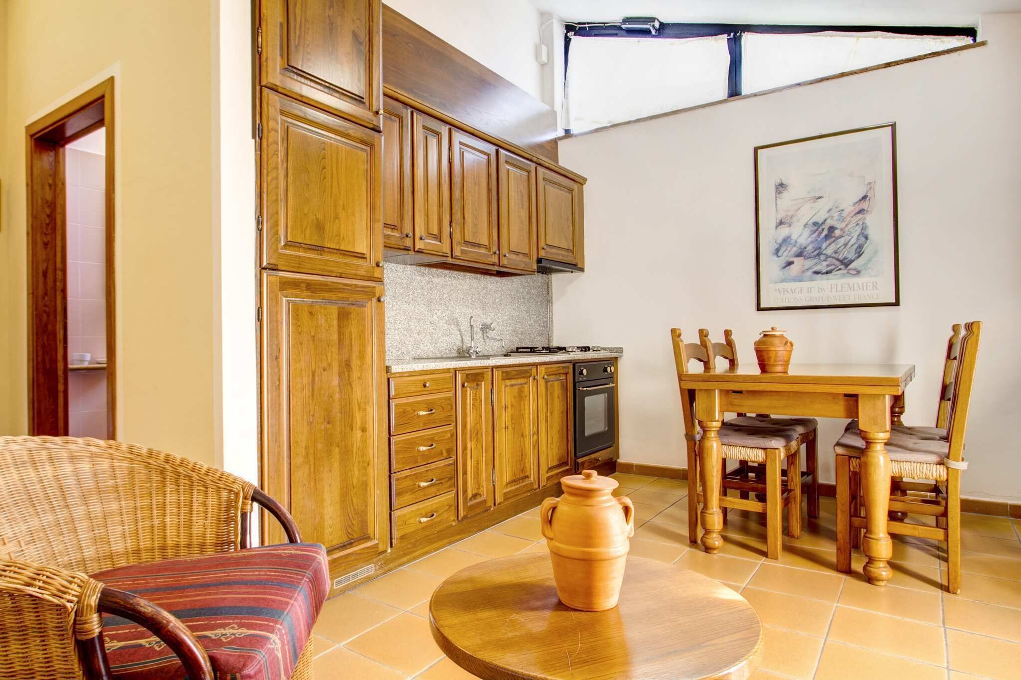Une cuisine avec des meubles en bois, une table à manger avec des chaises, un fauteuil en osier et une table ronde sur laquelle est posé un pot en argile.