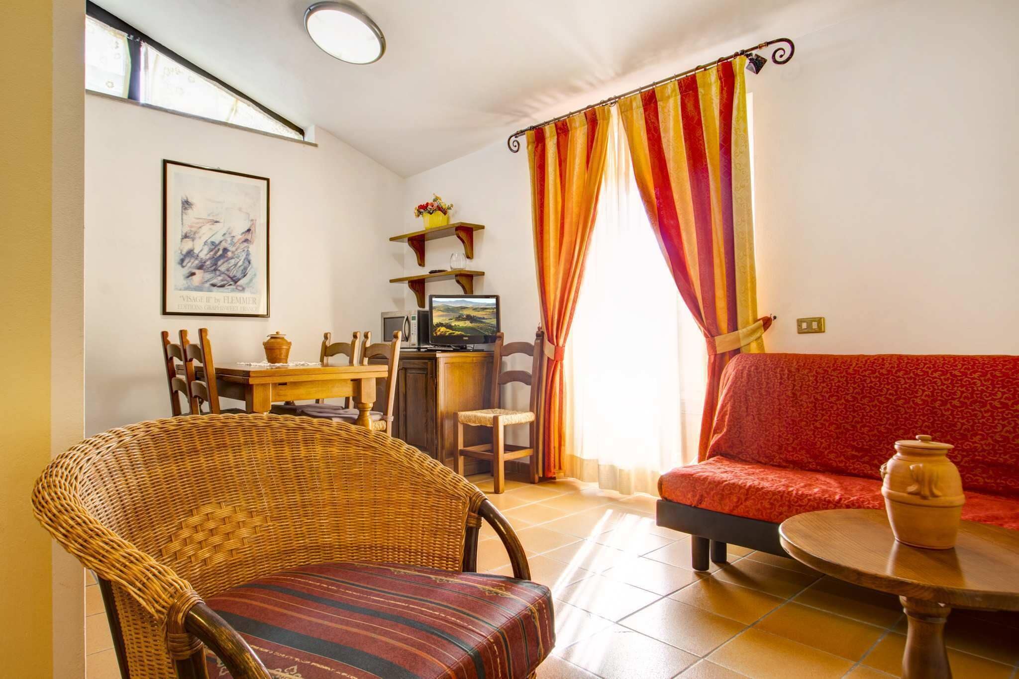 Una sala de estar con una silla de mimbre, un sofá rojo, una mesa de comedor con sillas, un televisor y una ventana con cortinas a rayas.