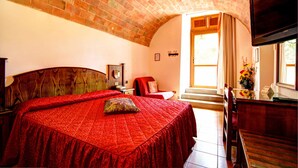 A bedroom with a brick ceiling, a large bed with a red bedspread, a wooden headboard, a TV mounted on the wall, and a window with curtains.