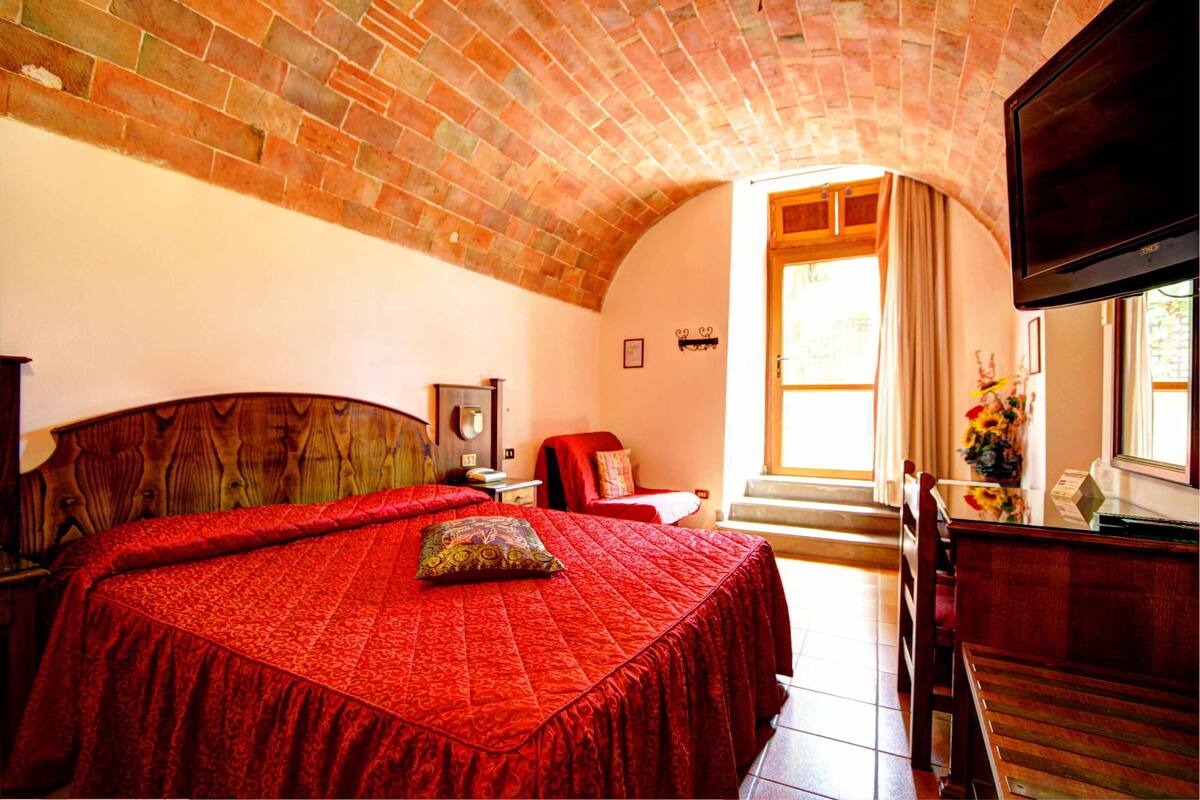 Une chambre avec un plafond en briques, un grand lit recouvert d’une couverture rouge, une tête de lit en bois, un téléviseur fixé au mur et une fenêtre avec des rideaux.