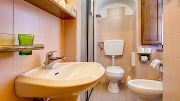 Ein Badezimmer mit Waschbecken, Toilette und Bidet, mit beigen Fliesen, einem Holzregal und einem grünen Glasobjekt an der Wand.