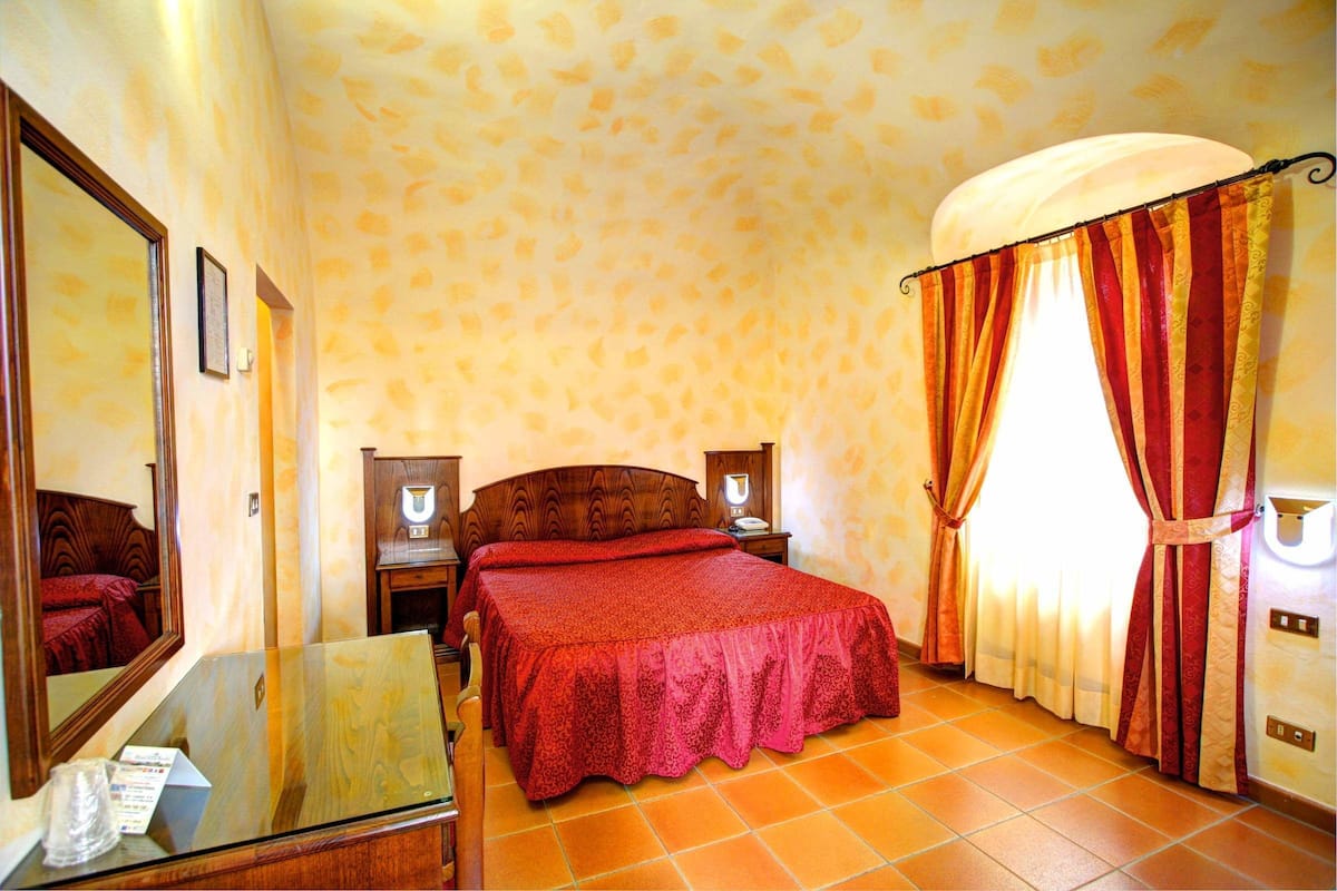 Une chambre d’hôtel avec un lit, une table de chevet, un miroir et une fenêtre avec des rideaux.