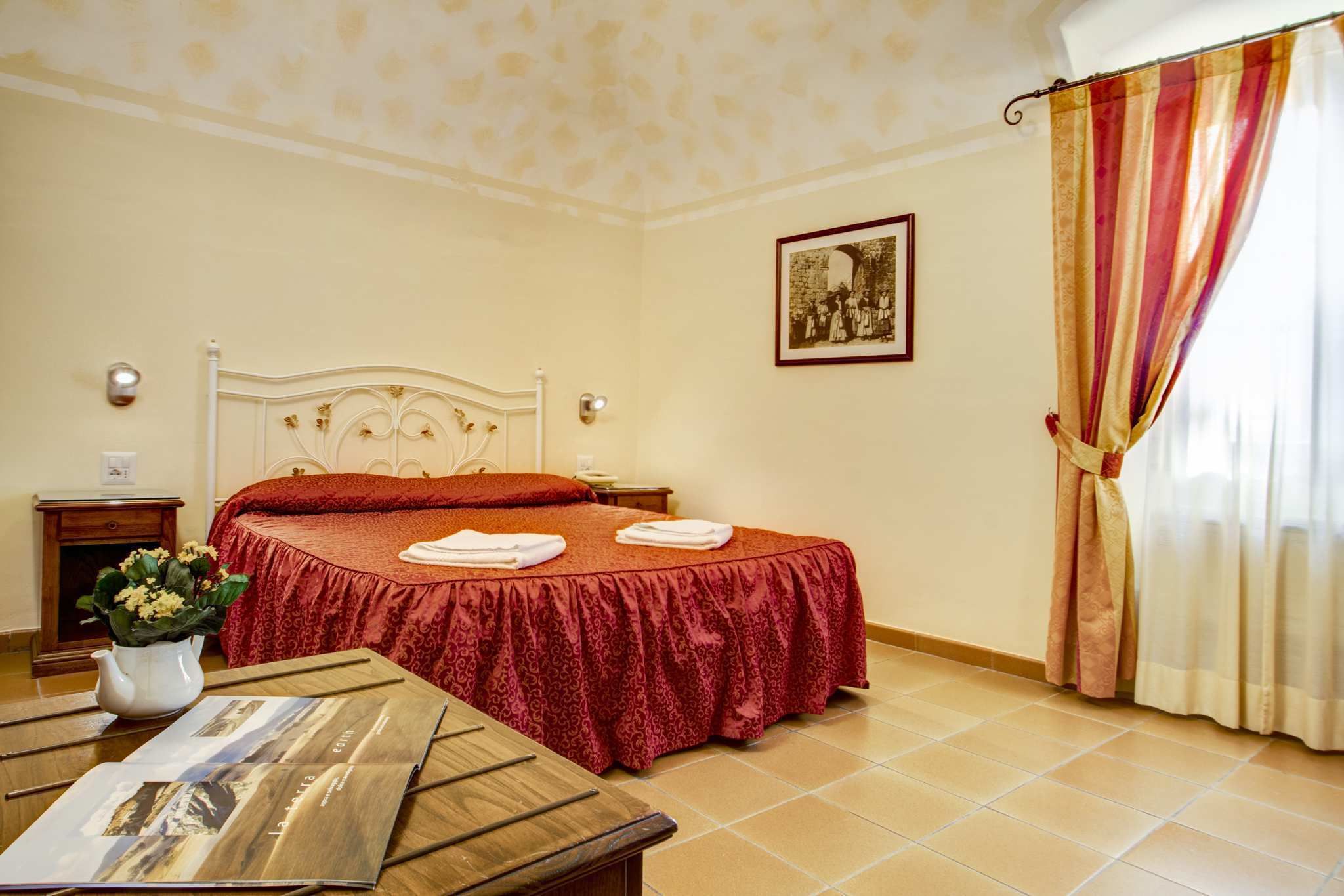 A hotel room with a bed, a nightstand, a table with a magazine, a vase with flowers, and a window with curtains.