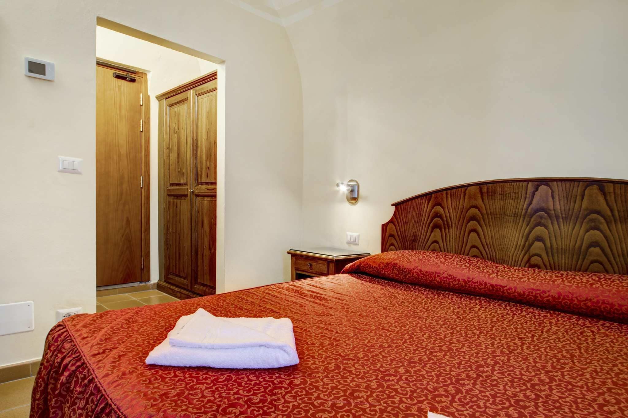 A hotel room with a bed, a wooden wardrobe, a nightstand, and a wall-mounted light fixture.
