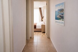 En gang der fører til et soveværelse med en seng, et natbord og et vindue med gardiner. Der er et indrammet billede af Antalya By på væggen.