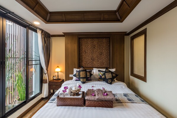 Una cama bien hecha con toallas y un cuenco decorativo, una mesita de noche con lámpara, y vistas al verdor por una ventana.