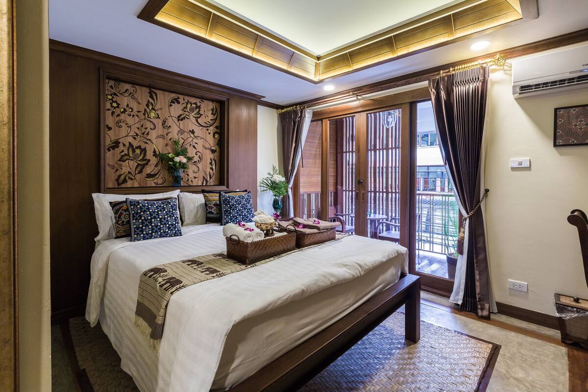 Habitación de hotel con una cama grande, cabecera de madera con detalles florales tallados, balcón con vistas y techo con iluminación empotrada.