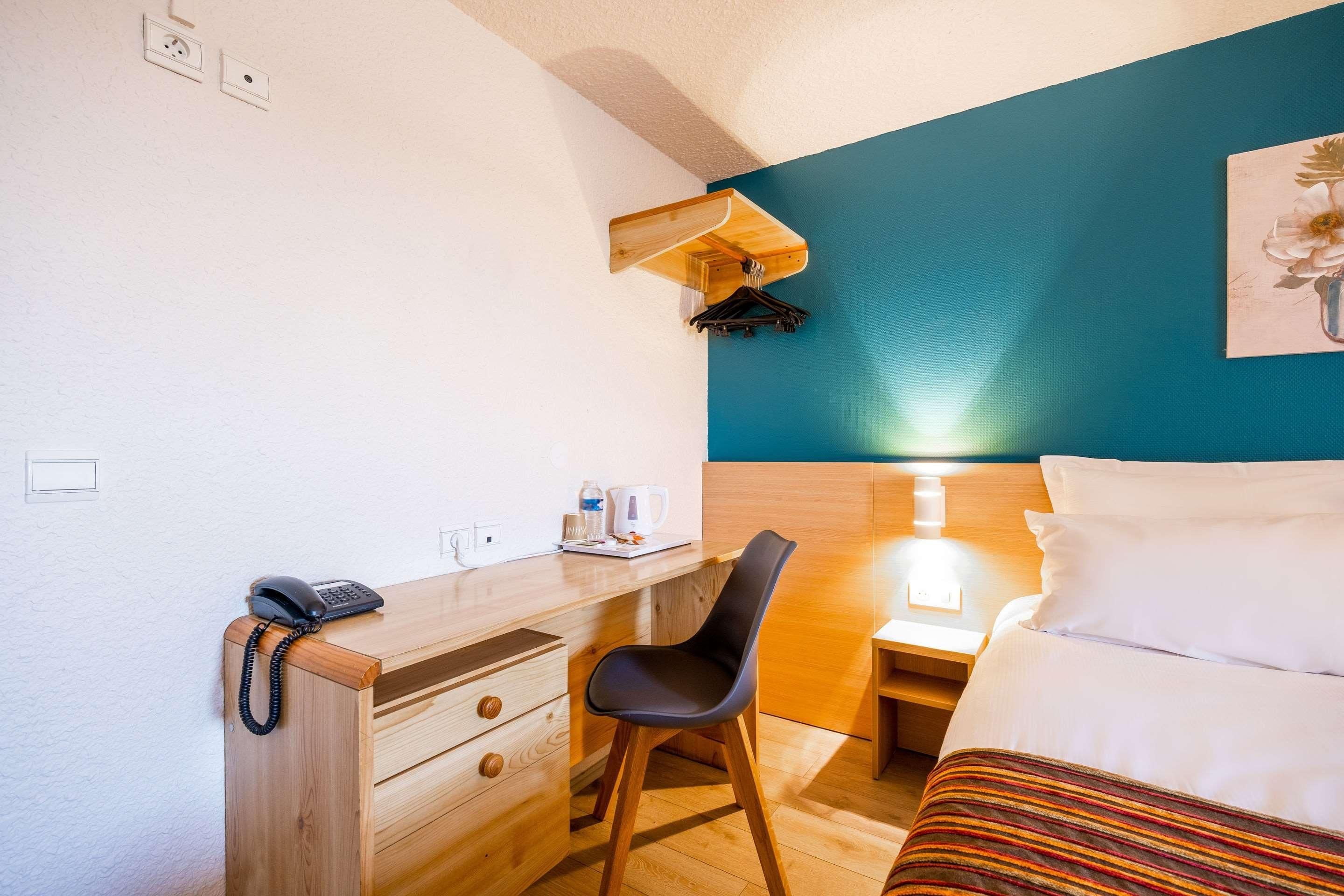 A hotel room with a bed, a desk with a telephone, a chair, a nightstand with a lamp, and a wall-mounted coat rack.