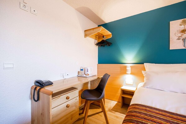 A hotel room with a bed, a desk with a telephone, a chair, a nightstand with a lamp, and a wall-mounted coat rack. - Comfort Hotel Montlucon (Montlucon)