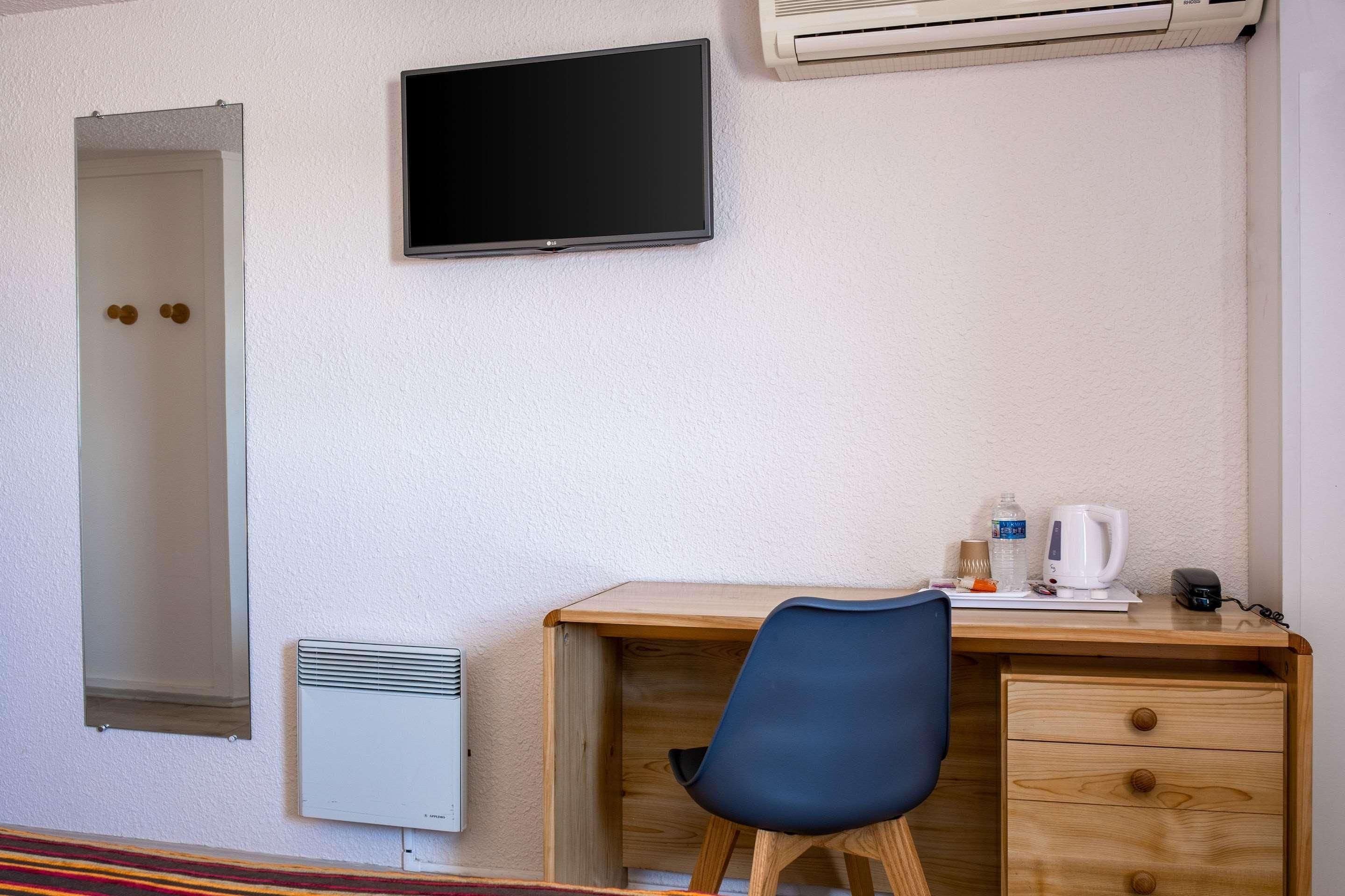 Habitación de hotel con un escritorio de madera, una silla, televisor montado en la pared, radiador y dispensador de agua.
