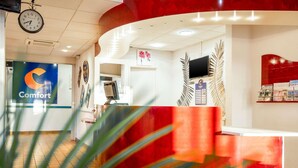 Un espace d’accueil comprenant une horloge, un panneau indiquant « Confort », un comptoir d’accueil, un téléviseur et un mur orné de tableaux.