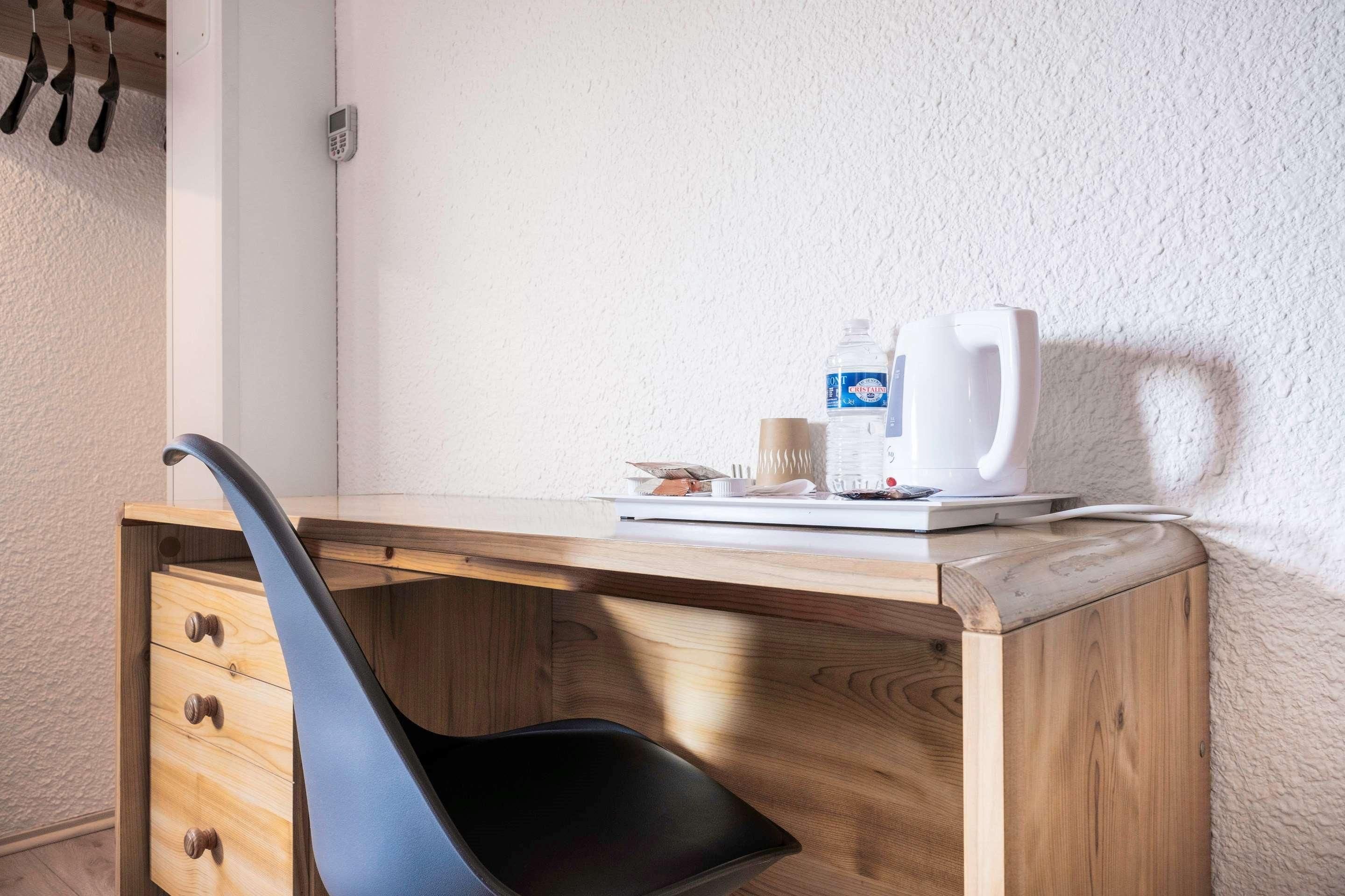 A wooden desk with a black chair, a white coffee maker, a bottle of water, and a cup.