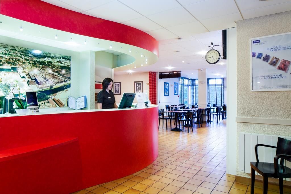 Zona de recepción de hotel con un mostrador rojo, un reloj y un área de comedor con mesas y sillas.