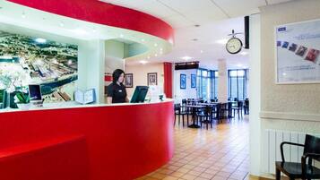 Zona de recepción de hotel con un mostrador rojo, un reloj y un área de comedor con mesas y sillas.