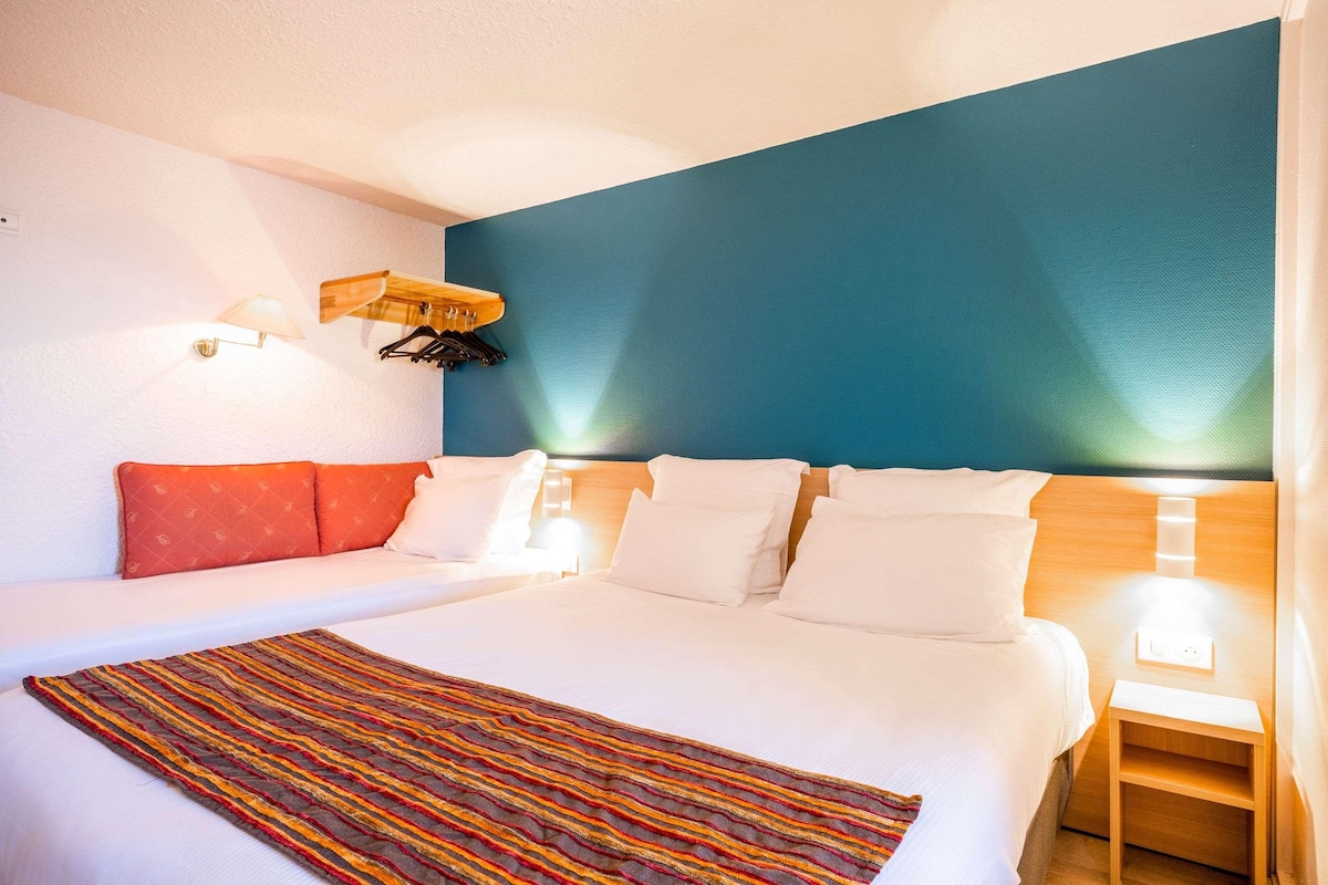 Une chambre d’hôtel avec un grand lit, du linge de lit blanc, un tapis rayé aux couleurs vives, une tête de lit en bois et un mur d’accent de couleur turquoise.