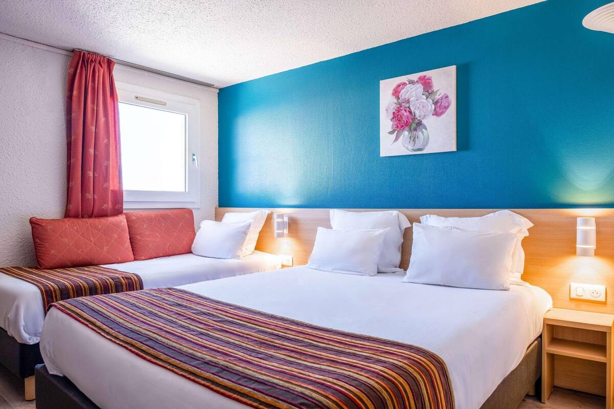 Une chambre d’hôtel avec deux lits, une grande fenêtre avec des rideaux rouges, un mur d’accent bleu et un tableau représentant des motifs floraux.