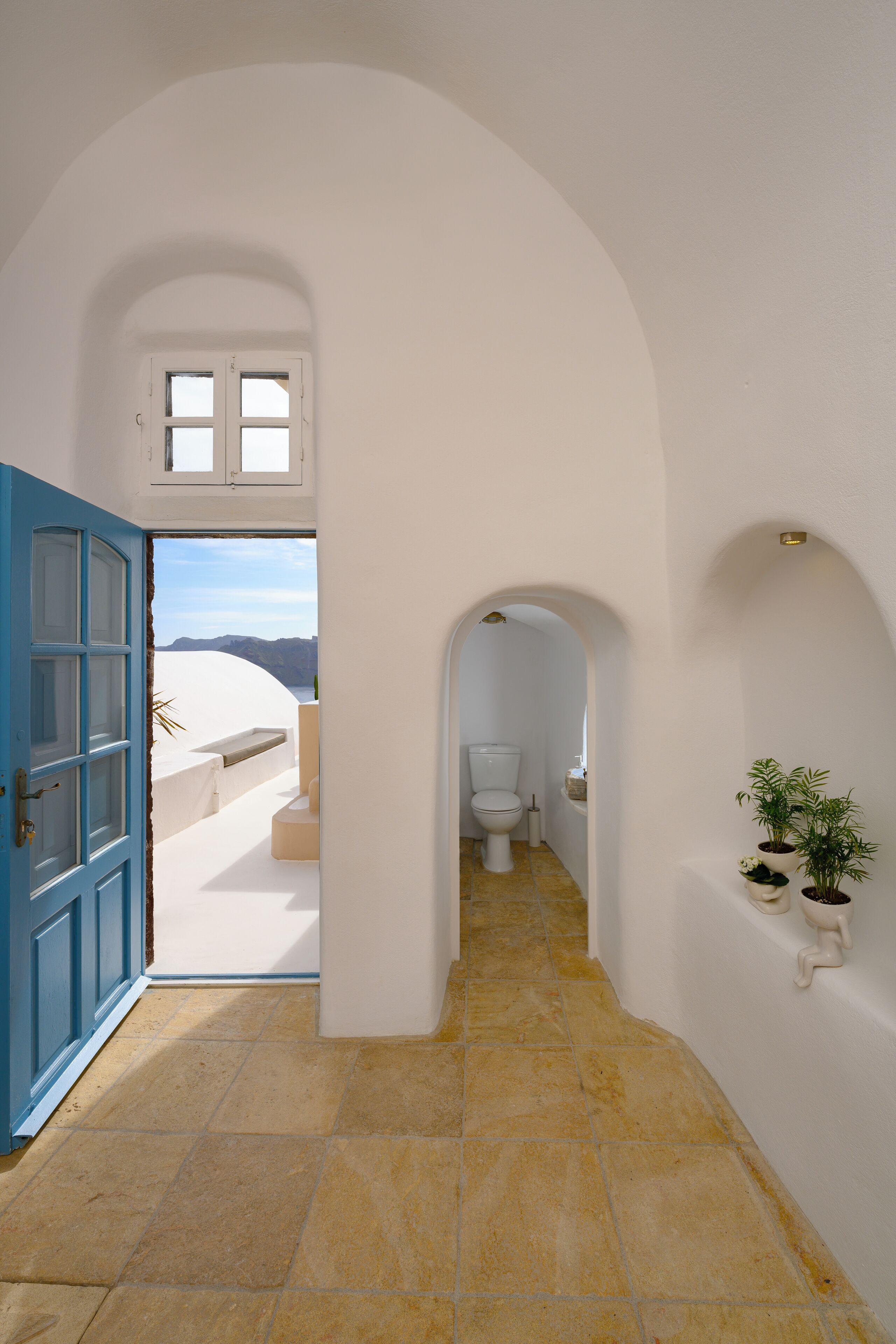 Un couloir avec une porte bleue donnant sur un balcon, des toilettes blanches et des plantes en pot.