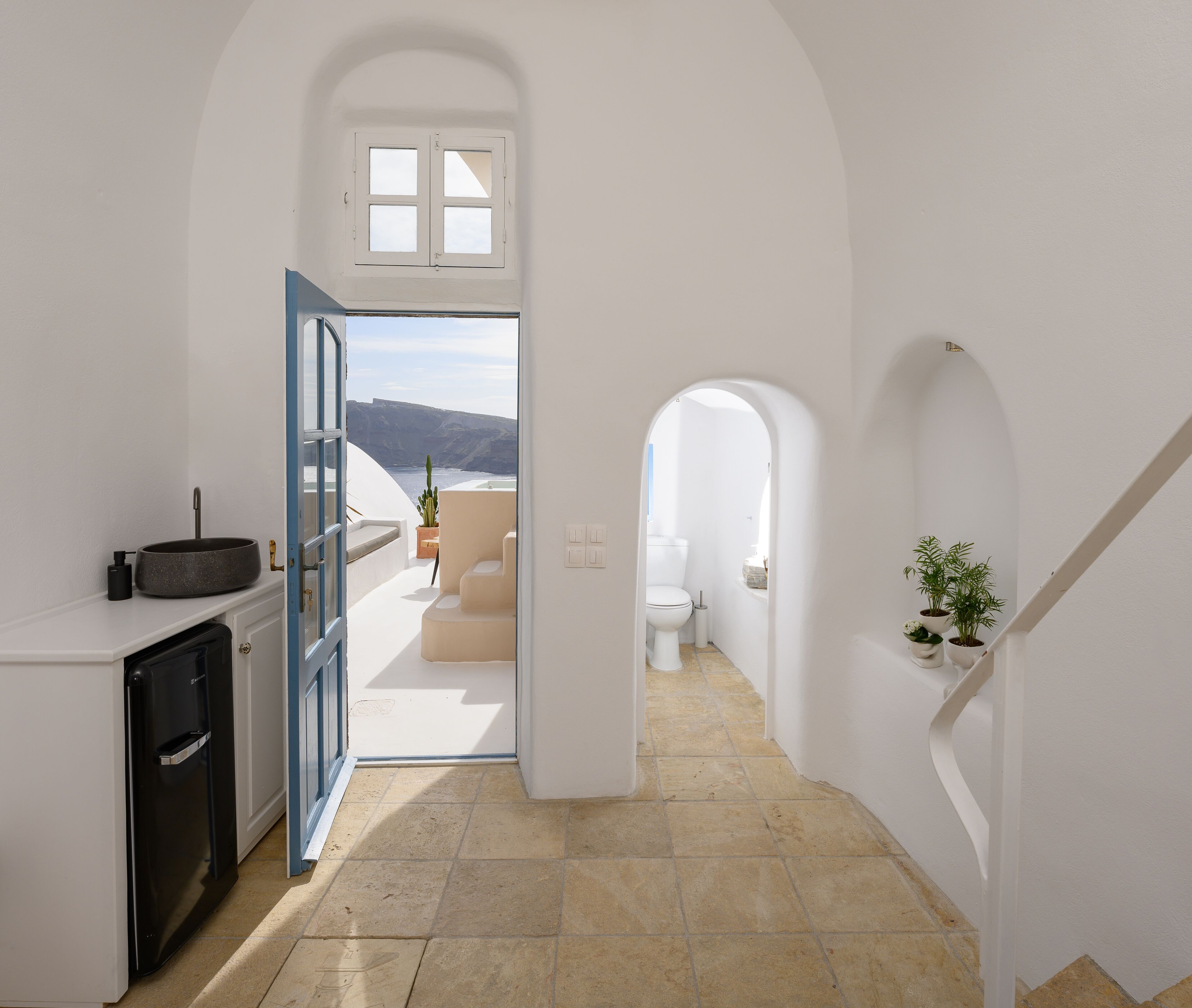 Un pasillo estrecho con una zona de cocina a la izquierda, un baño con inodoro y lavamanos, y vista al mar a través de una puerta abierta.