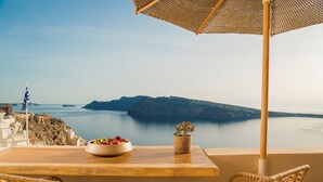 A table with wicker chairs and a woven umbrella overlooking a coastal view with a bowl of fruit and a potted plant.