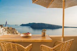 A table with wicker chairs and a woven umbrella overlooking a coastal view with a bowl of fruit and a potted plant.