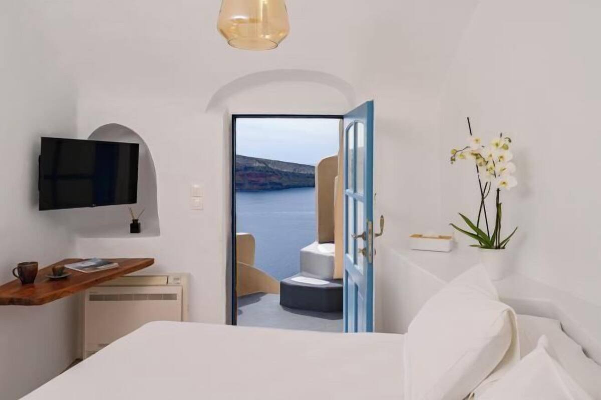Un dormitorio con cama, televisor, escritorio con una taza, planta y vista al mar a través de una puerta abierta.