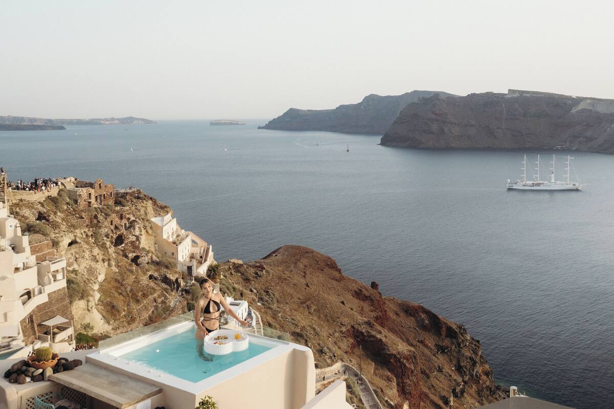 Un hotel en un acantilado con piscina y vistas al mar, un barco a lo lejos y terreno rocoso.