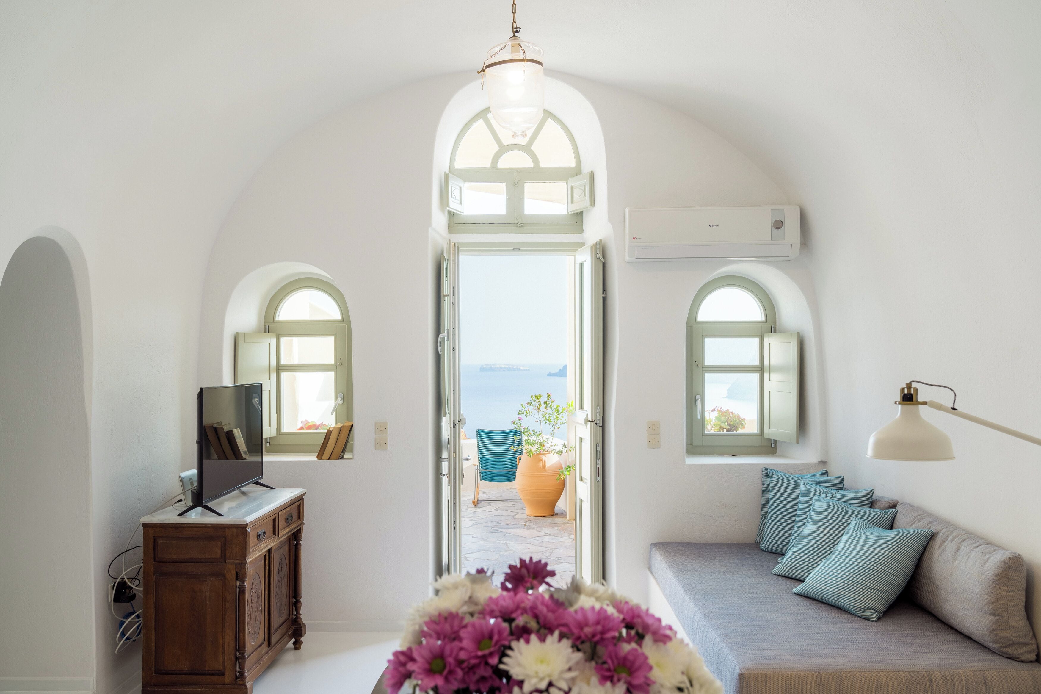 A room with a television on a wooden cabinet, a sofa with pillows, a potted plant, and a view through an arched doorway.