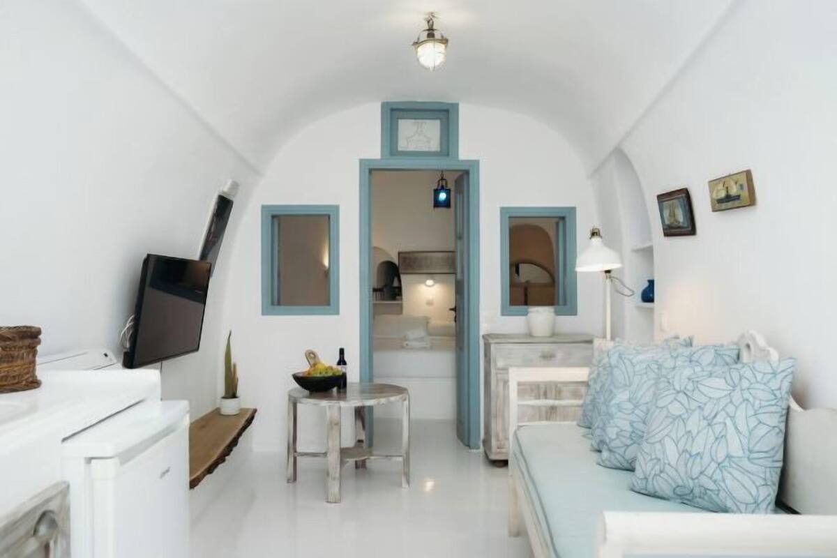 Un espacio habitable compacto con un televisor, una mesita con un cuenco, un sofá con cojines estampados, y una cocina visible a través de un arco.